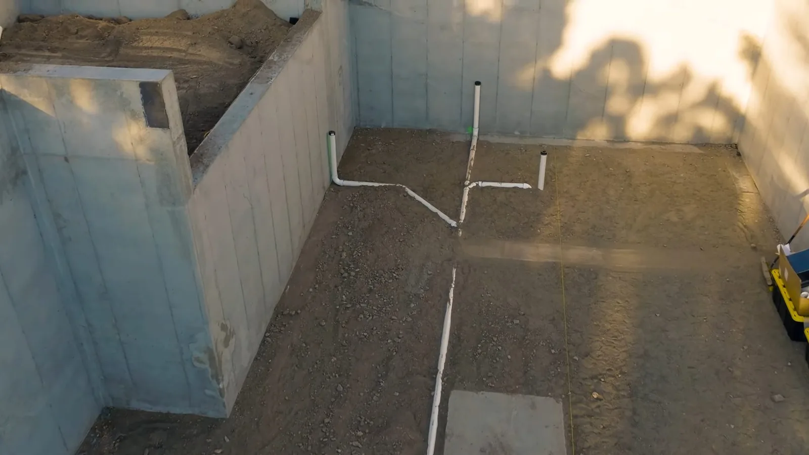 Excavated basement floor with white drain pipes laid in the soil and a wooden form along one side