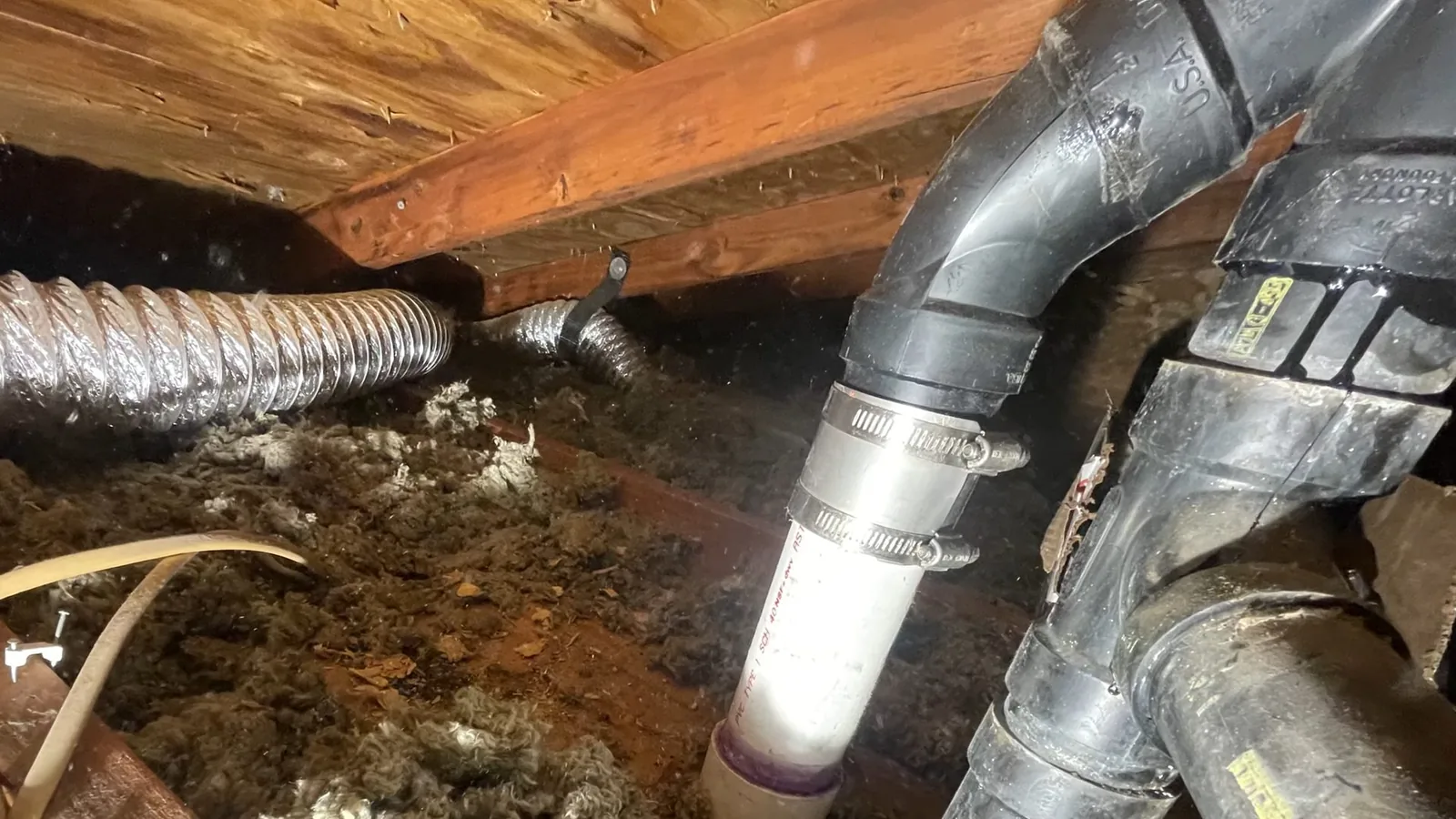 Black and white drain pipes joined with steel-clamped couplings under wood floor joists, above bare ground with insulation scraps