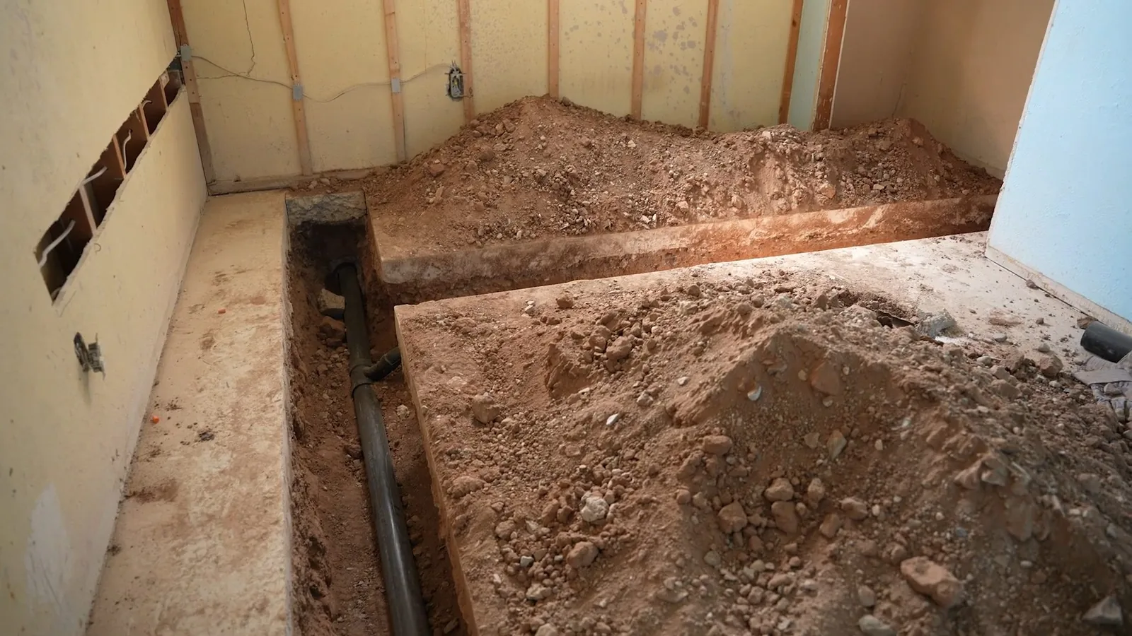 Basement room with the floor dug open along a drain pipe, piles of soil on both sides and bare wall framing behind
