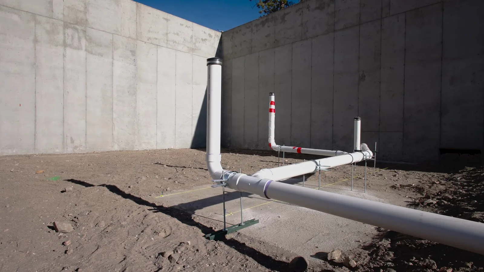 White plastic drain pipes rising from bare soil beside a tall concrete wall, with a marker post beside the run