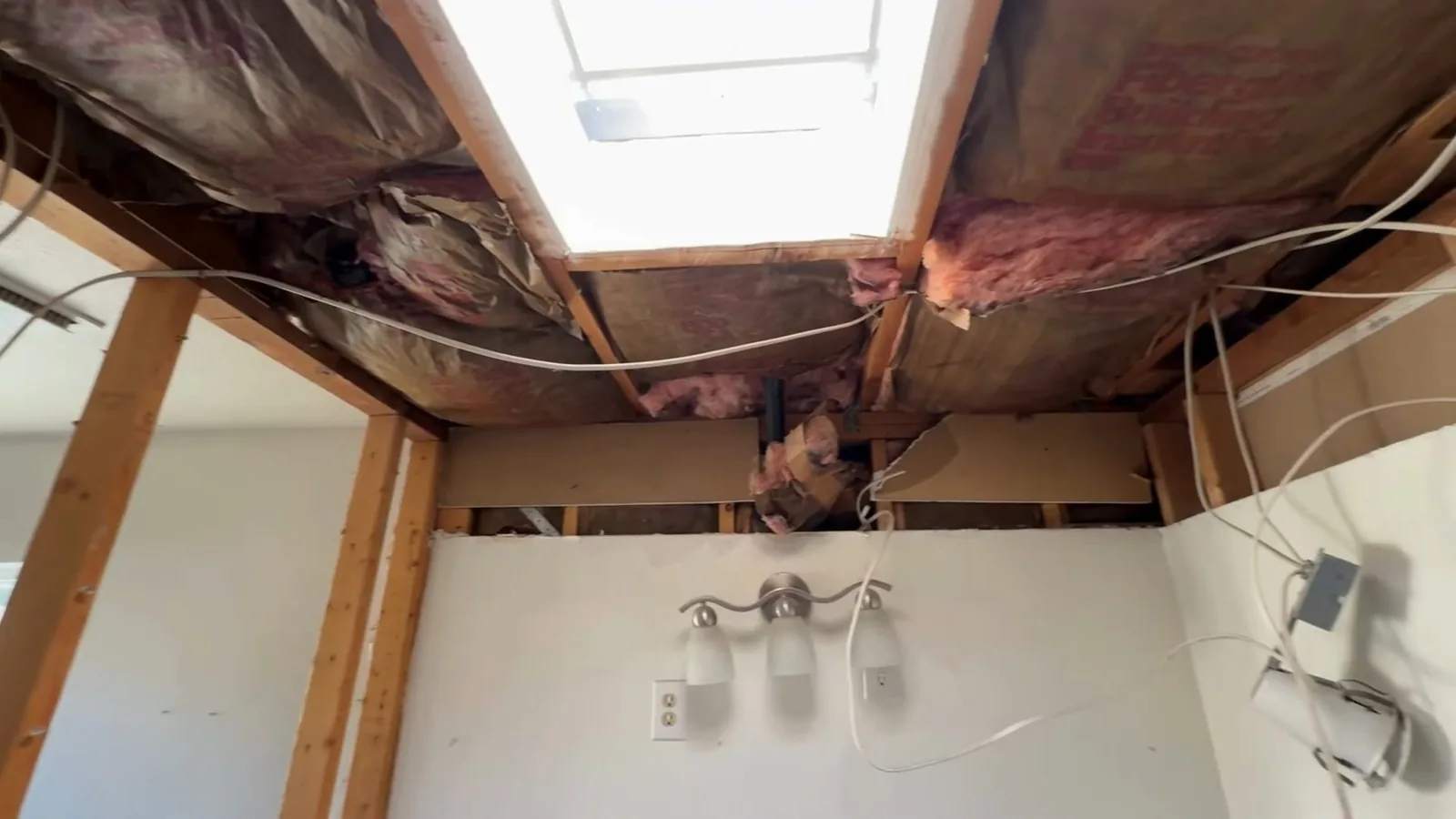 Bathroom ceiling cut open to show wood joists and pink insulation around a skylight shaft, above a wall with a light fixture