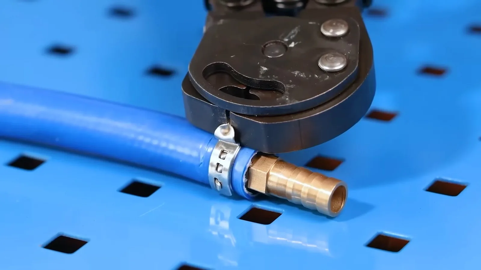 Crimp tool closed around a blue flexible pipe fitted with a brass connector, on a blue perforated board