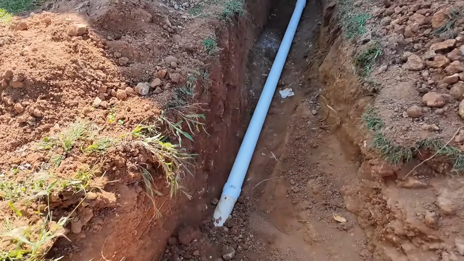 Narrow trench in red clay soil with a white pipe lying along the bottom and grass at the edges
