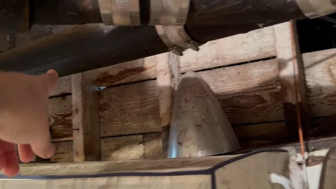 Large cast-iron drain pipe with steel band couplings running below wood floor joists, with a hand reaching in at the left