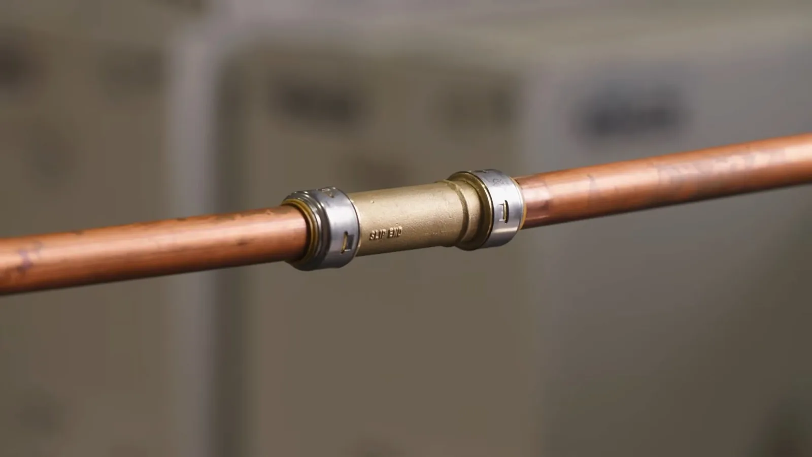 Horizontal copper pipe joined by a brass and steel push-fit coupling, against a blurred indoor background
