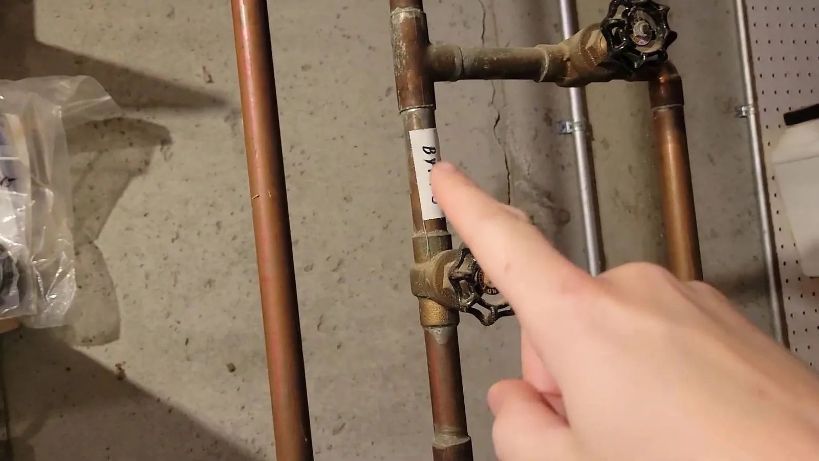 Hand pointing to a brass gate valve on a copper pipe run along a concrete basement wall