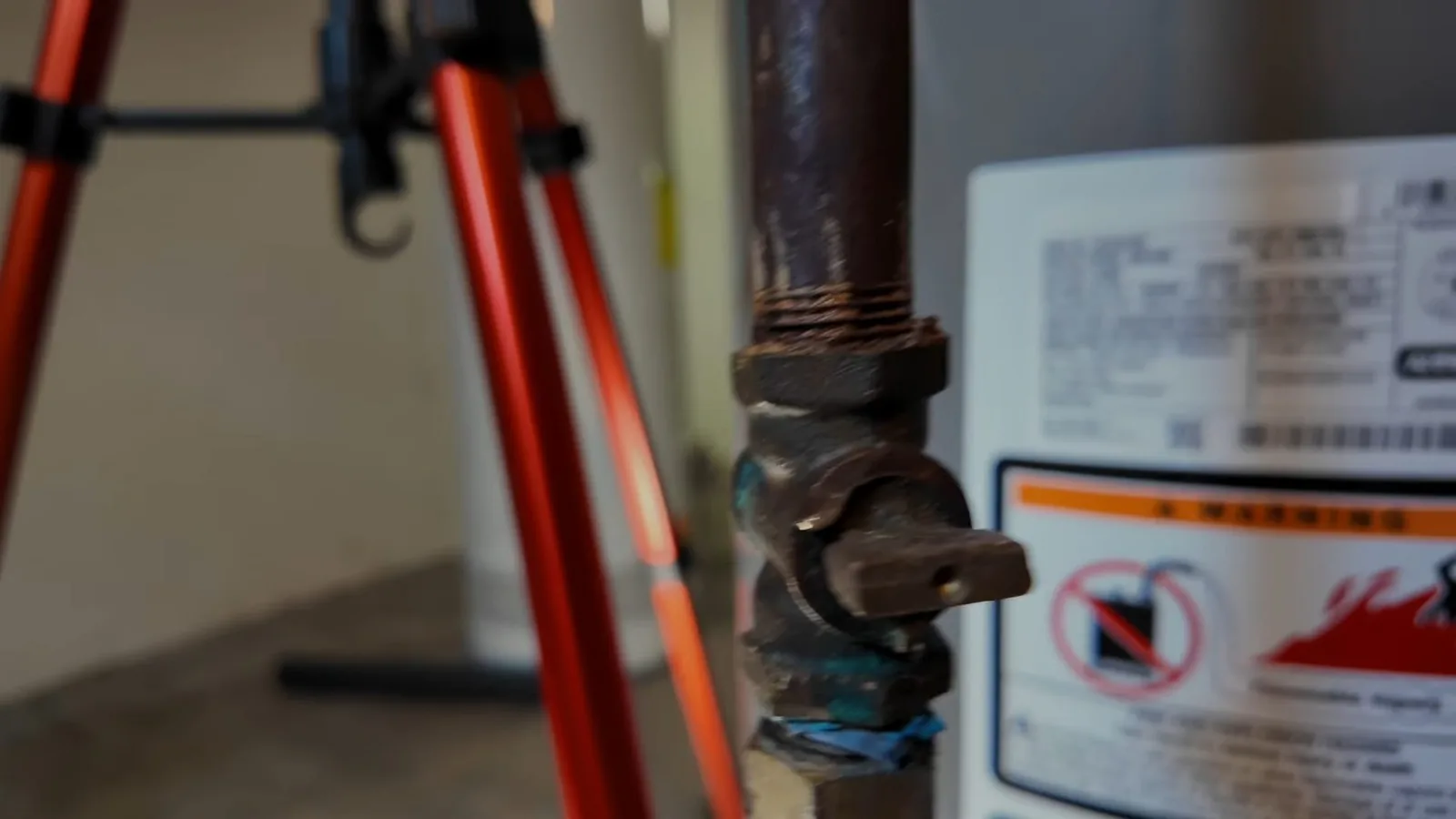 Corroded gas shutoff valve on a black iron pipe beside a water heater with a warning label, and a red tripod stand behind