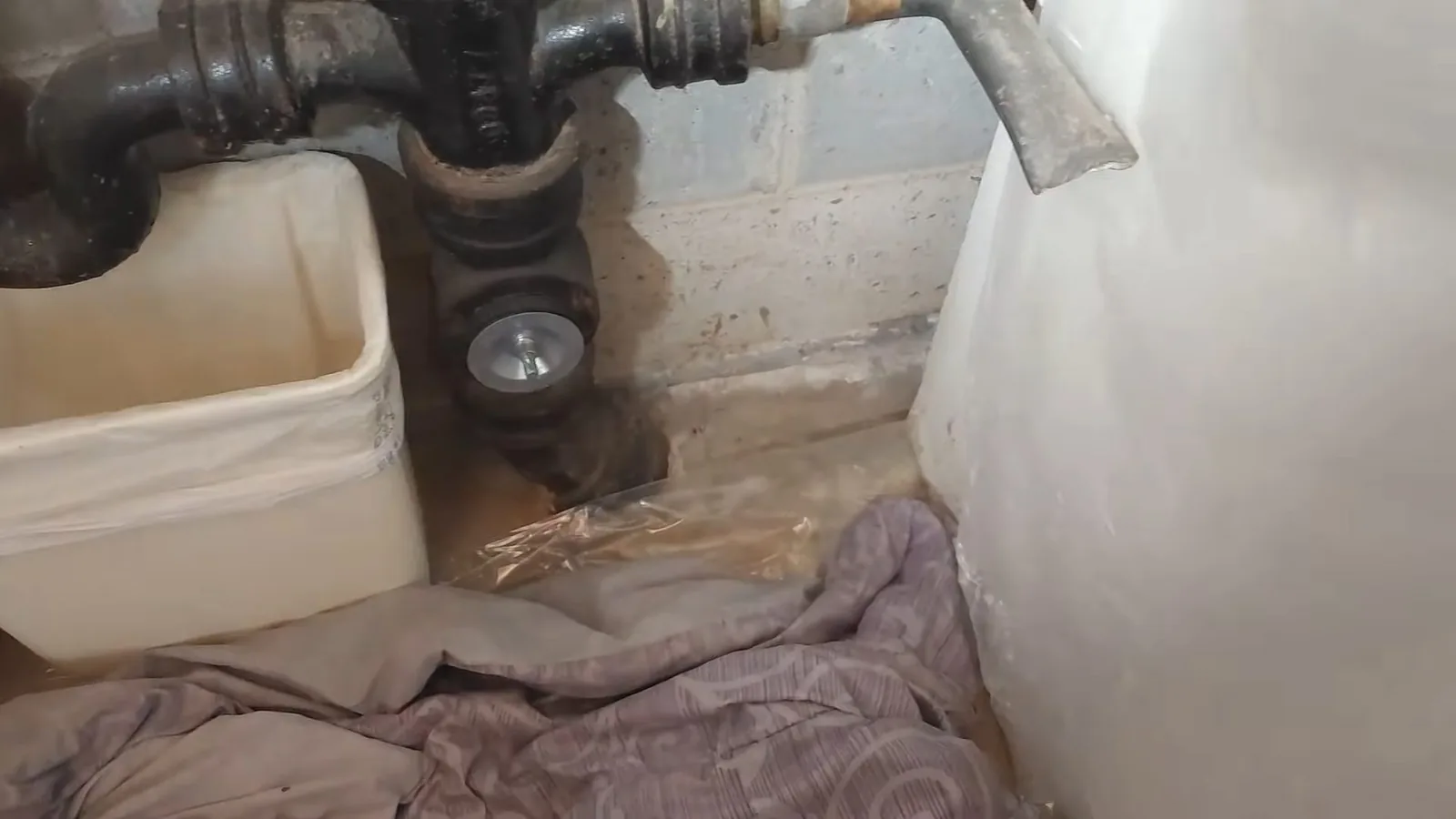 Old black cast-iron drain fittings next to a stained floor drain and a concrete block wall