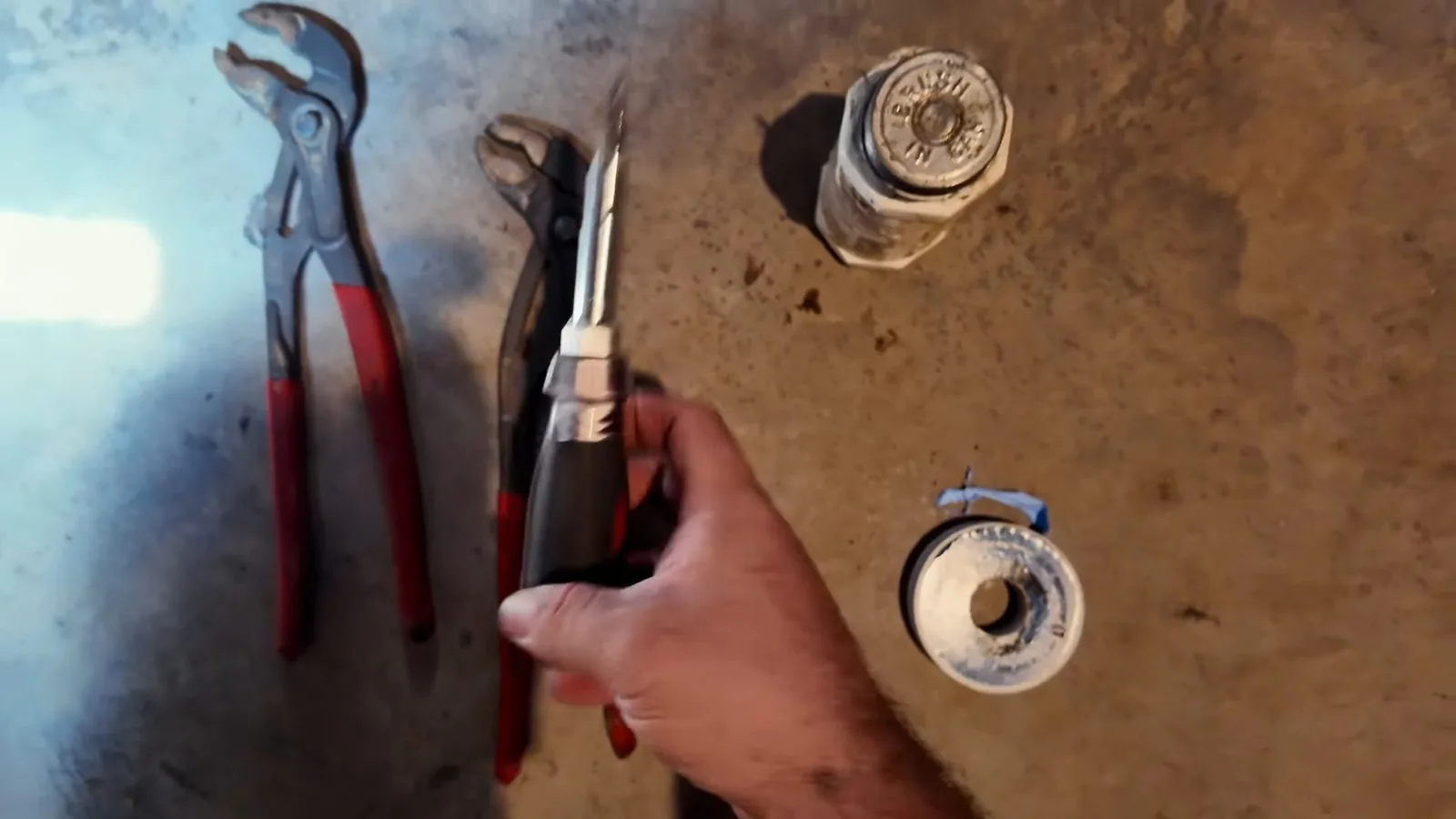 Hand holding red-handled pliers on a stained floor beside a roll of thread tape and a threaded fitting