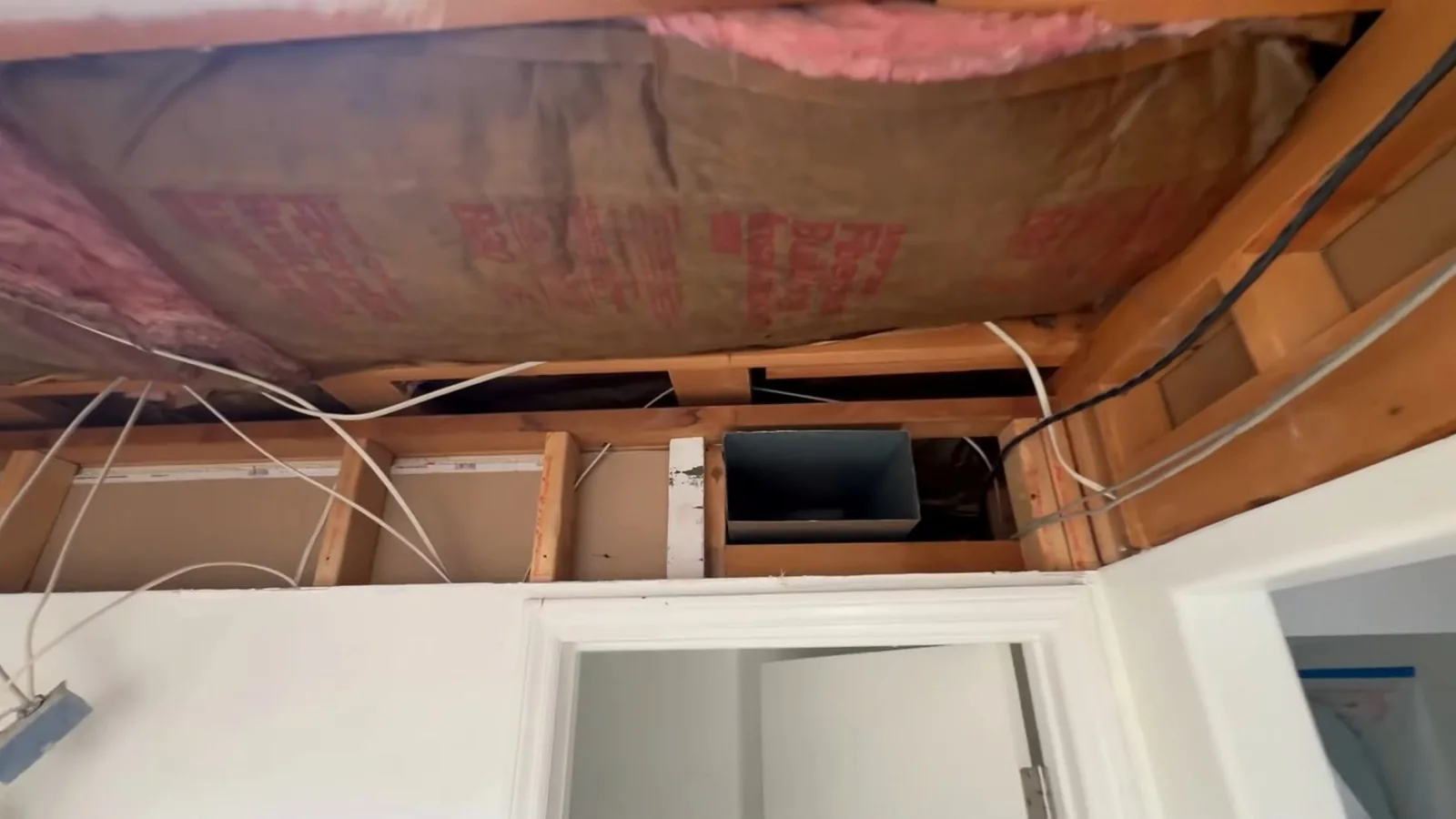 Bathroom ceiling and shower wall opened up, with pink insulation, wiring and framing exposed above a white wall