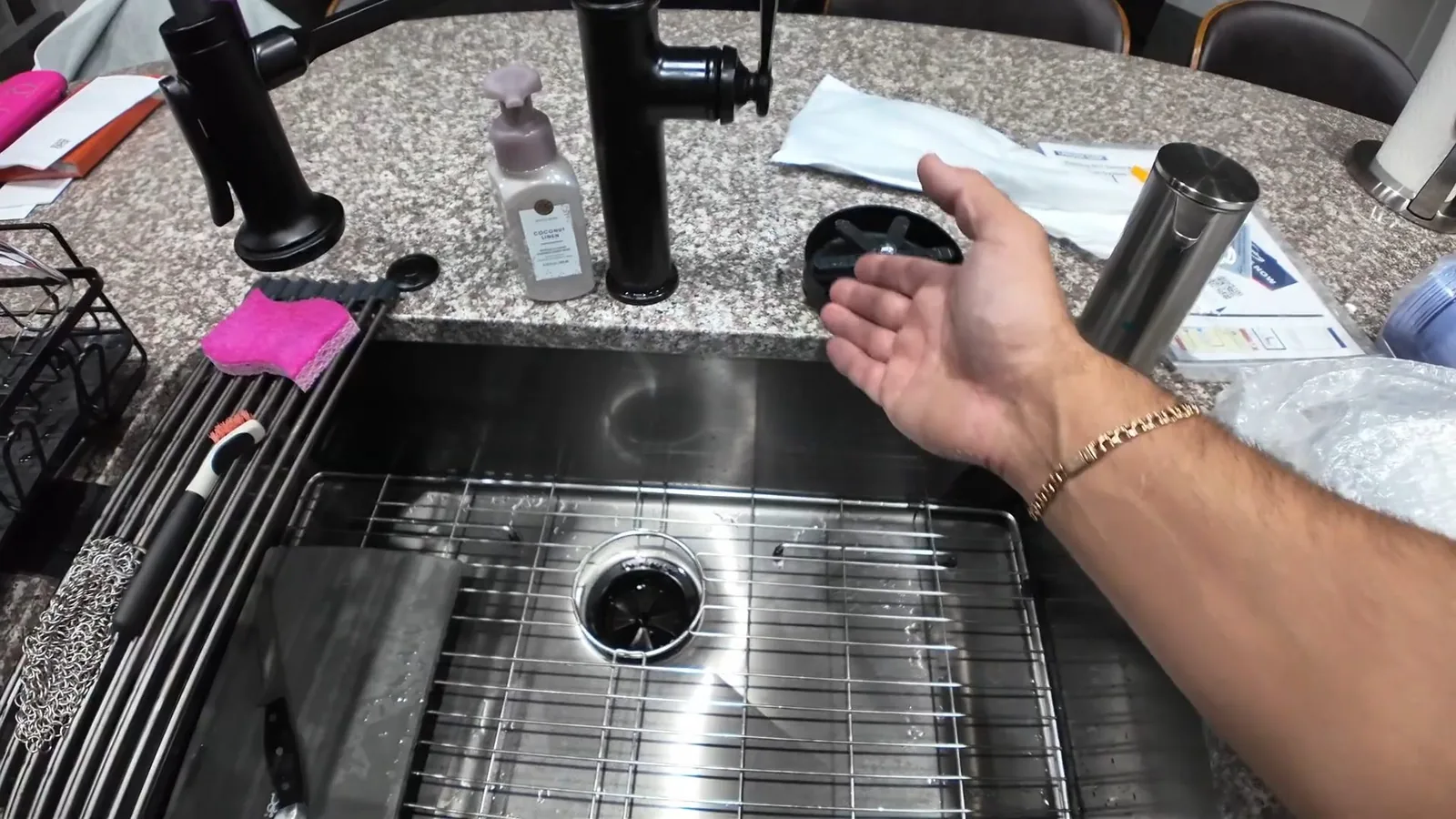 Forearm reaching over a stainless steel kitchen sink with a black faucet, a soap bottle and small parts laid out on the granite counter