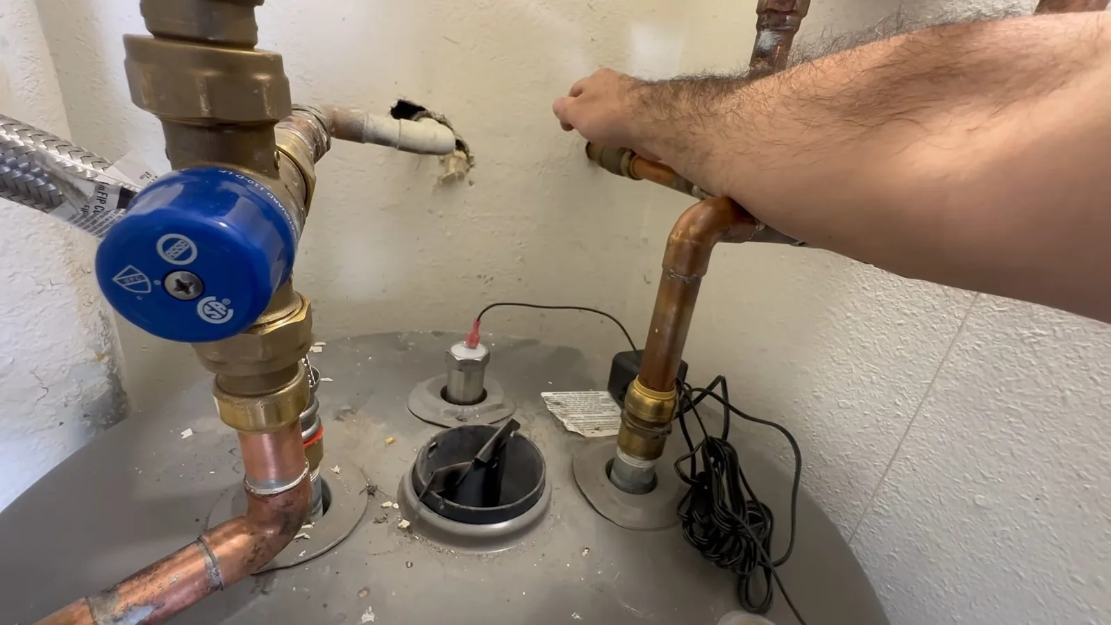 A forearm reaching across the top of a tank water heater, with a blue-handled shutoff valve, copper supply pipes and a flexible hose connected above it