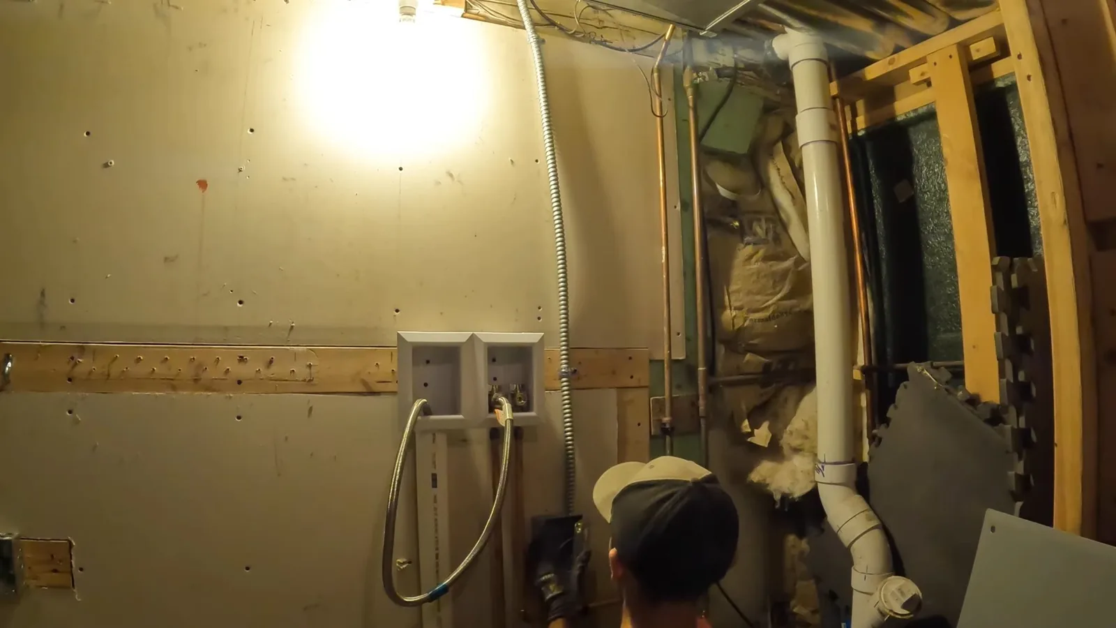 Person kneeling at a framed basement wall with a white drain pipe and a laundry supply box beside it