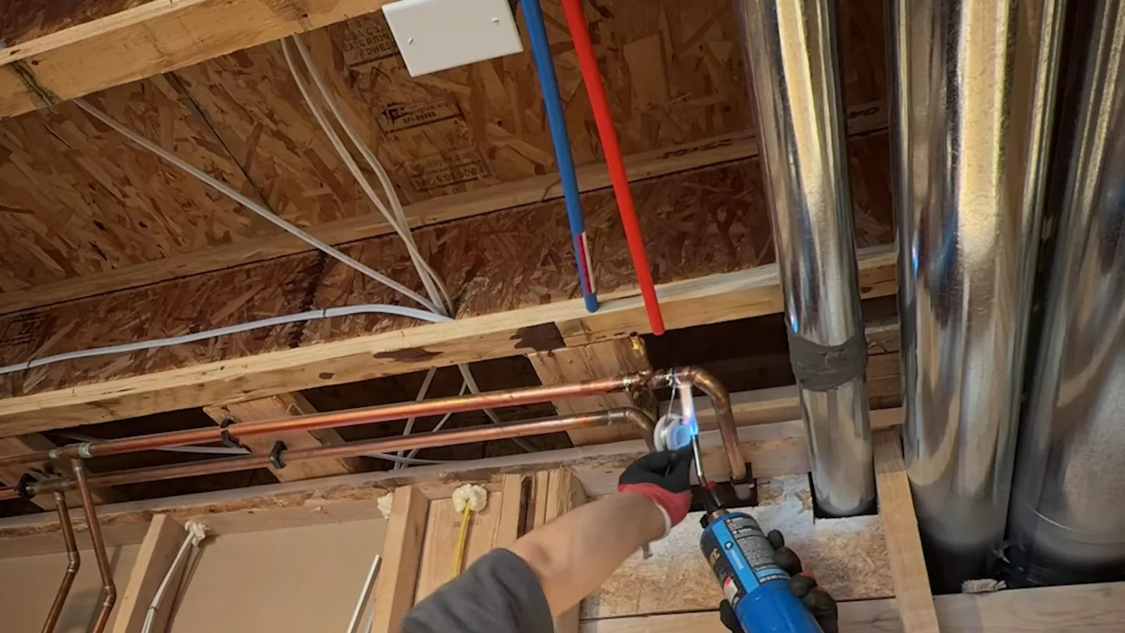 Gloved hand with a blue torch soldering a copper joint under a framed basement ceiling with red and blue PEX lines