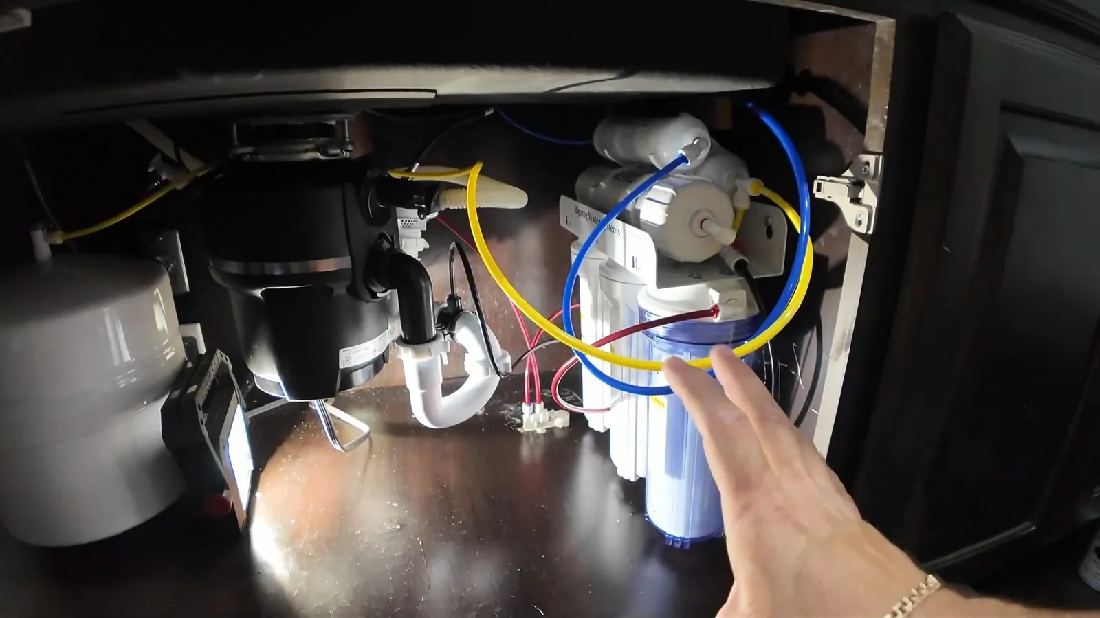 Open sink cabinet showing a garbage disposal with a white drain trap, and a multi-stage water filter with blue and yellow tubing, beside a pointing hand