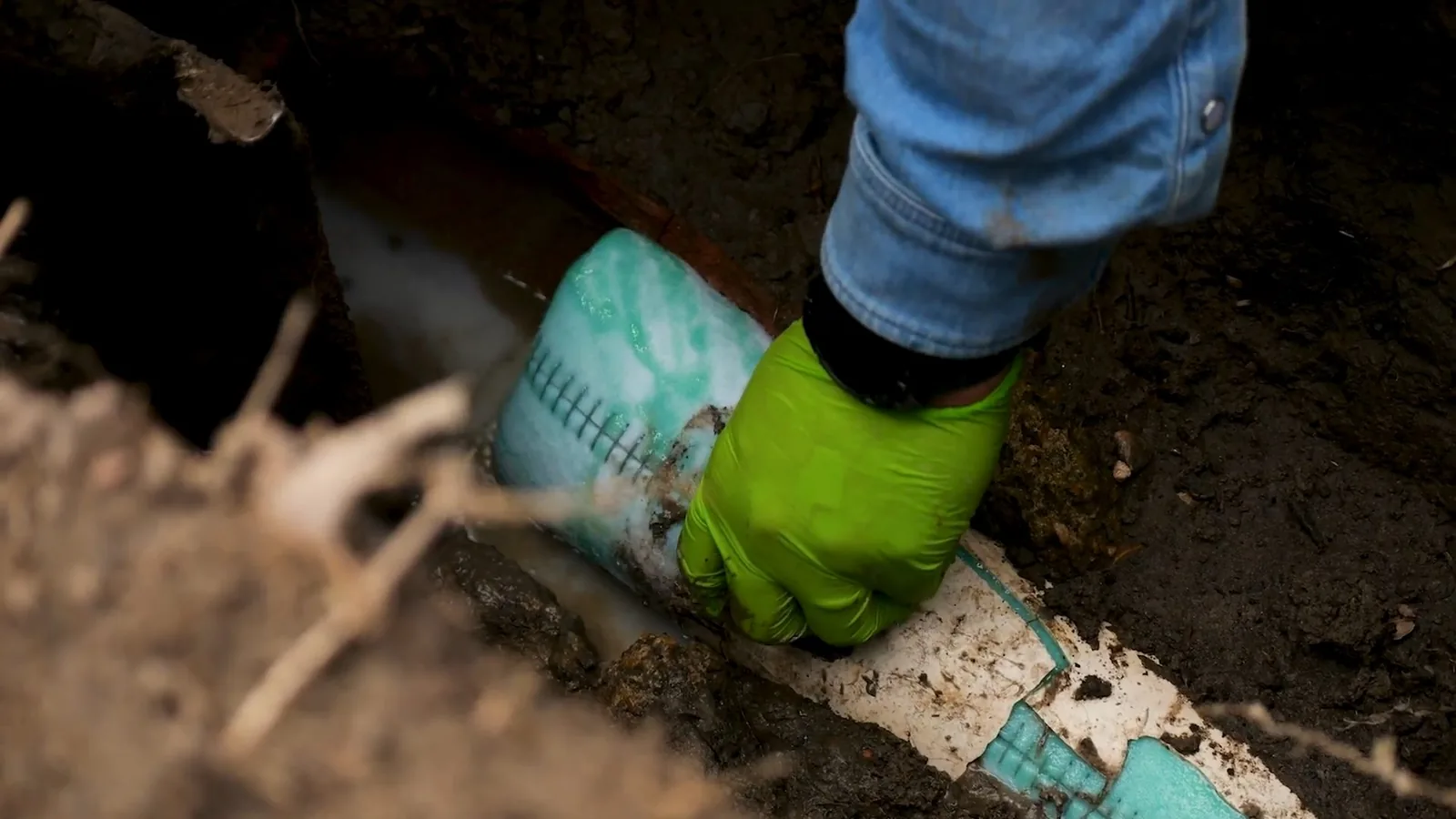 Green-gloved hand guiding a teal and white pipe liner into an old pipe in an excavated pit