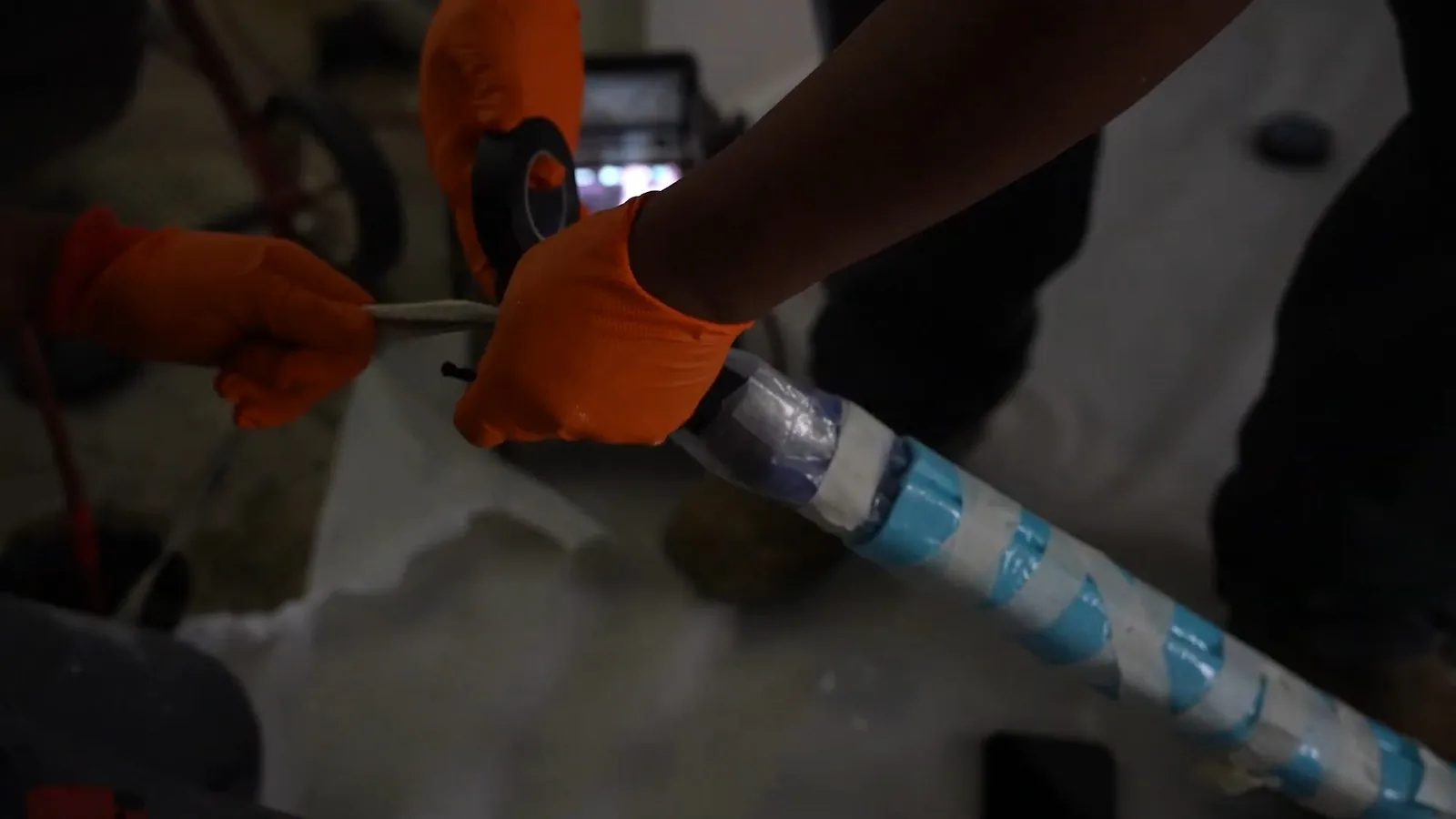 Orange-gloved hands wrapping blue tape around a liner tube while another gloved hand holds it steady