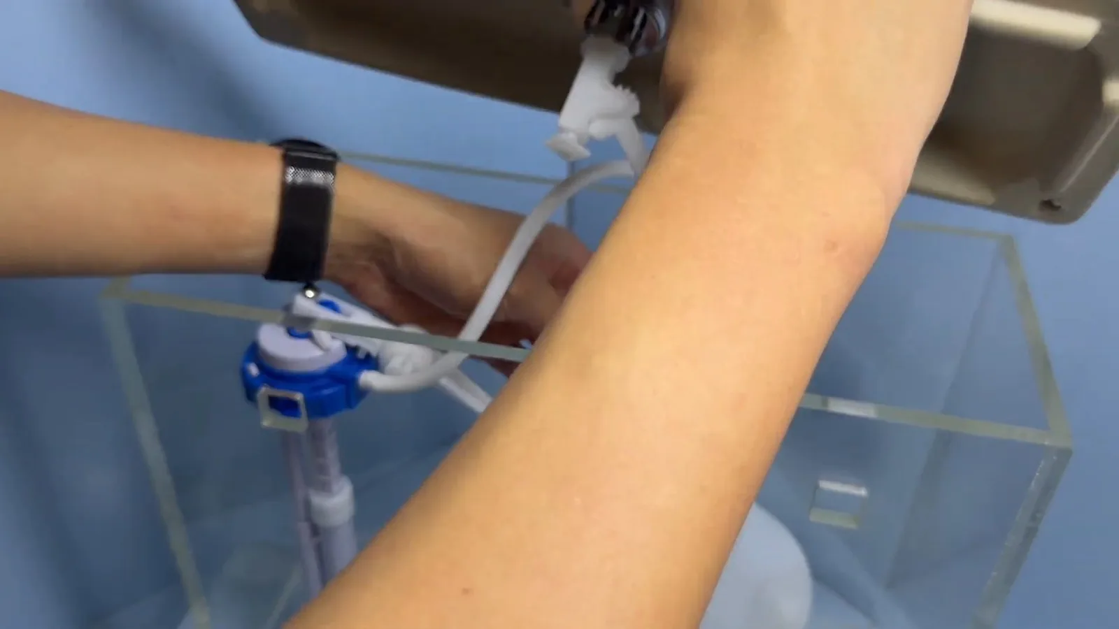 Forearm reaching into a clear acrylic demonstration toilet tank to adjust a blue and white fill valve
