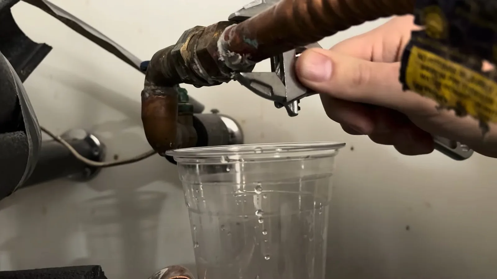 Hand using a wrench to loosen a fitting on a water heater inlet line, with a clear plastic cup below catching drips