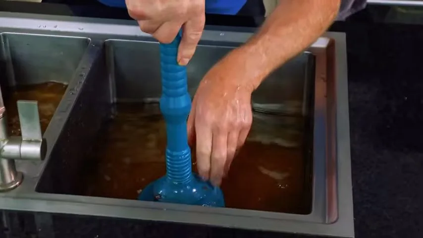 Hand pushing a blue plunger up and down in a stainless steel kitchen sink filled with brown water