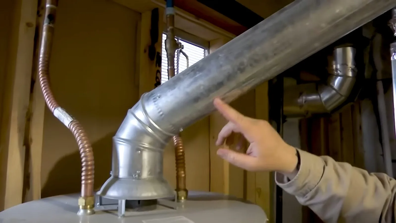 Hand pointing to a galvanized vent elbow above a water heater in an unfinished basement, with copper lines on both sides
