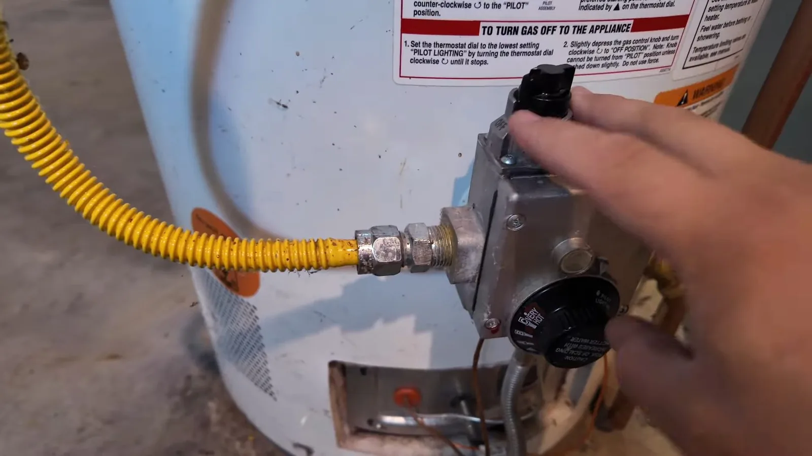 Hand turning the gas control knob on a tank water heater, with a yellow flexible gas line connected and an instruction label above