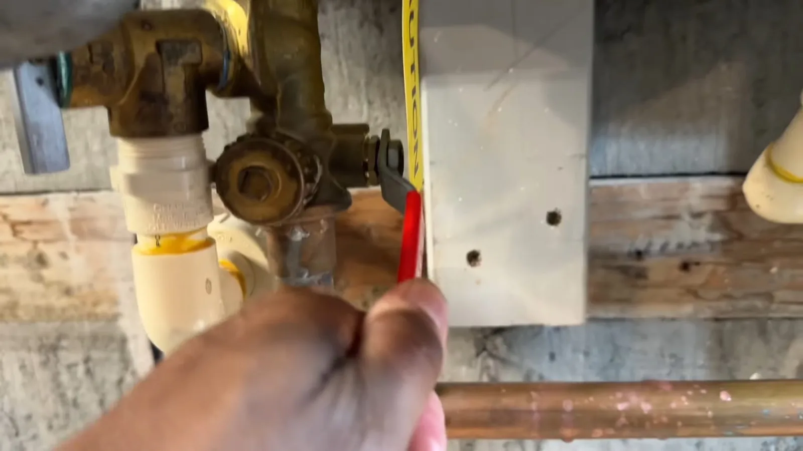 Hand turning a red-handled service valve on copper and brass pipes under a tankless water heater, with a yellow tape measure beside it