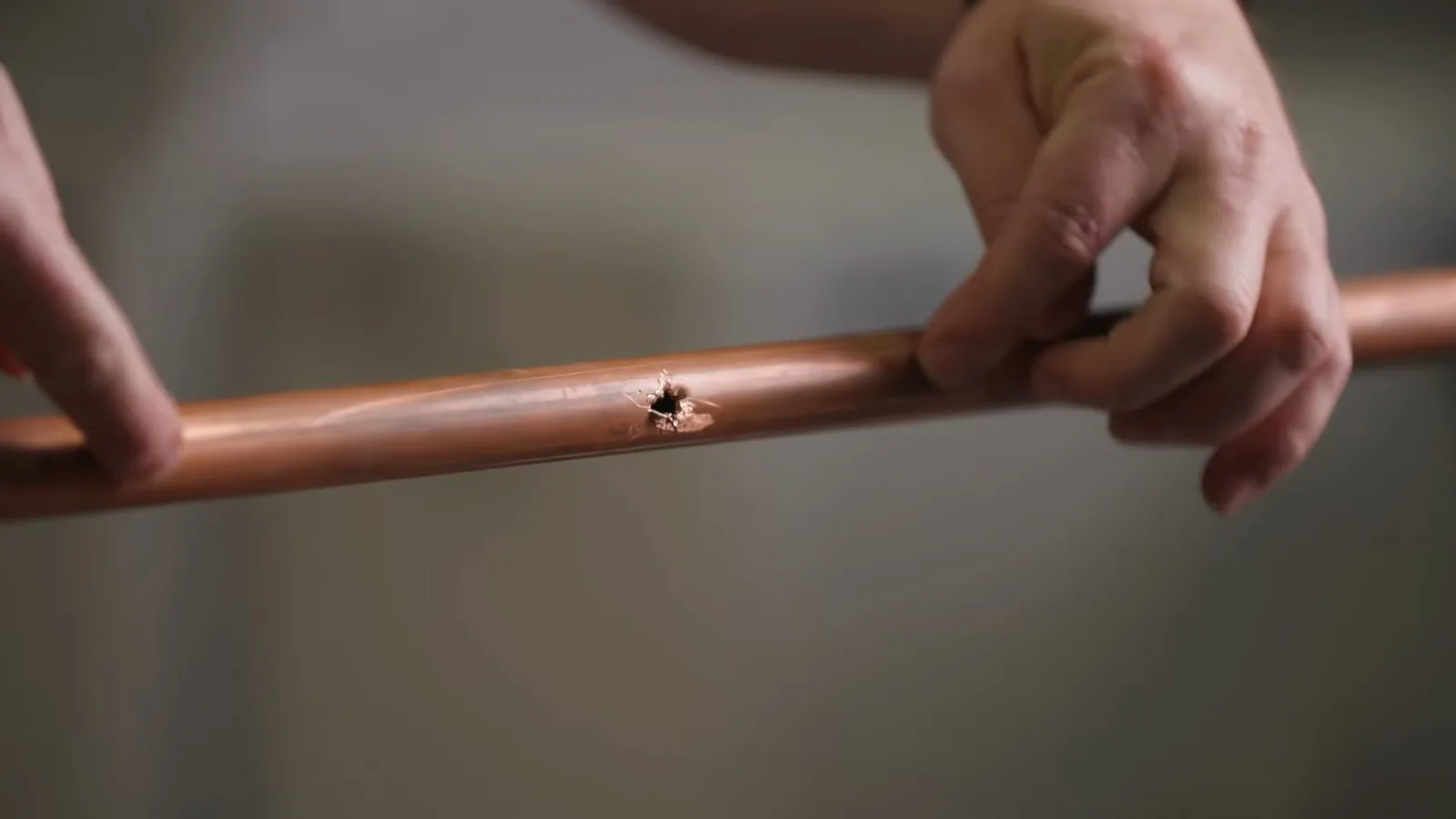Two hands holding a length of copper pipe that has a small pinhole puncture in its side