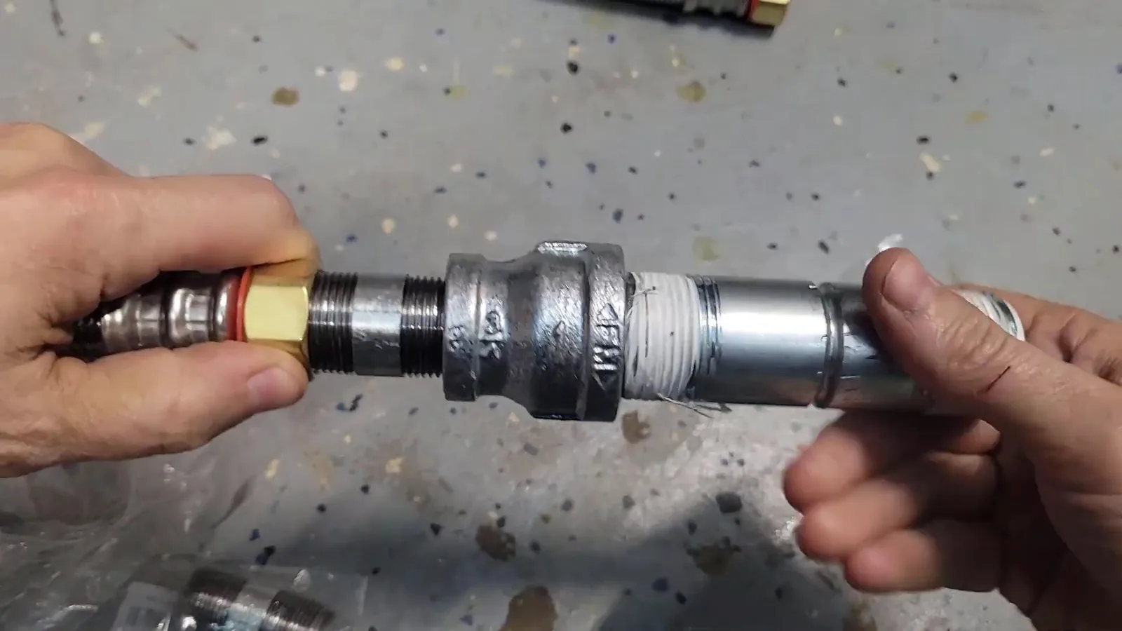 Two hands holding a black iron pipe coupling joined to threaded steel pipe and a brass fitting, over a speckled garage floor
