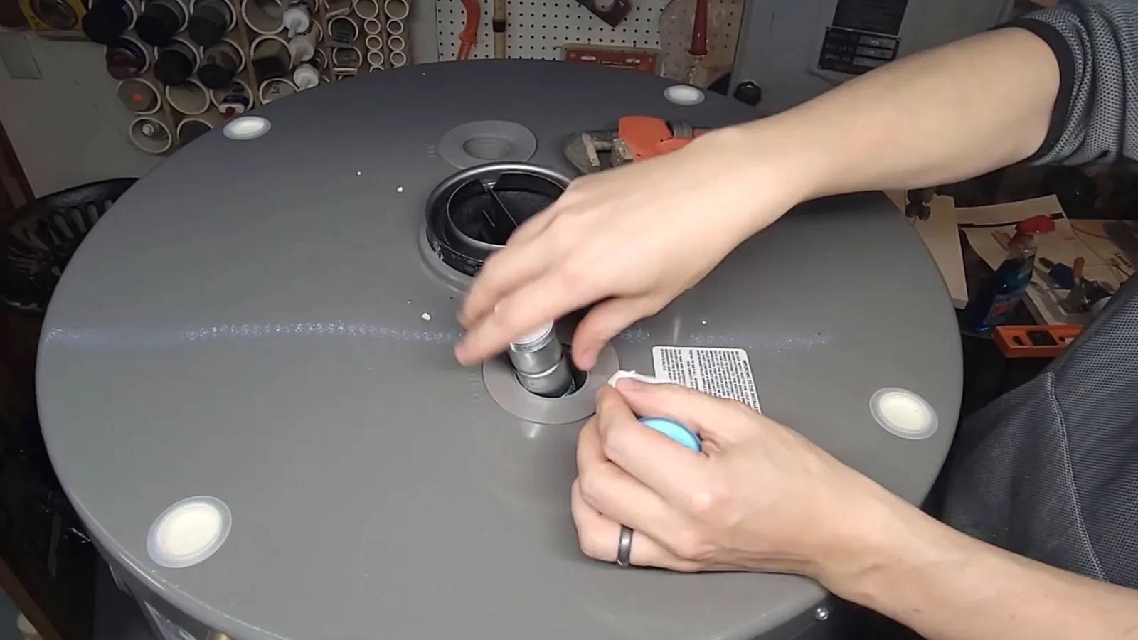 Two hands tightening a threaded fitting on the gray top of a tank water heater, in a workshop with pegboard behind