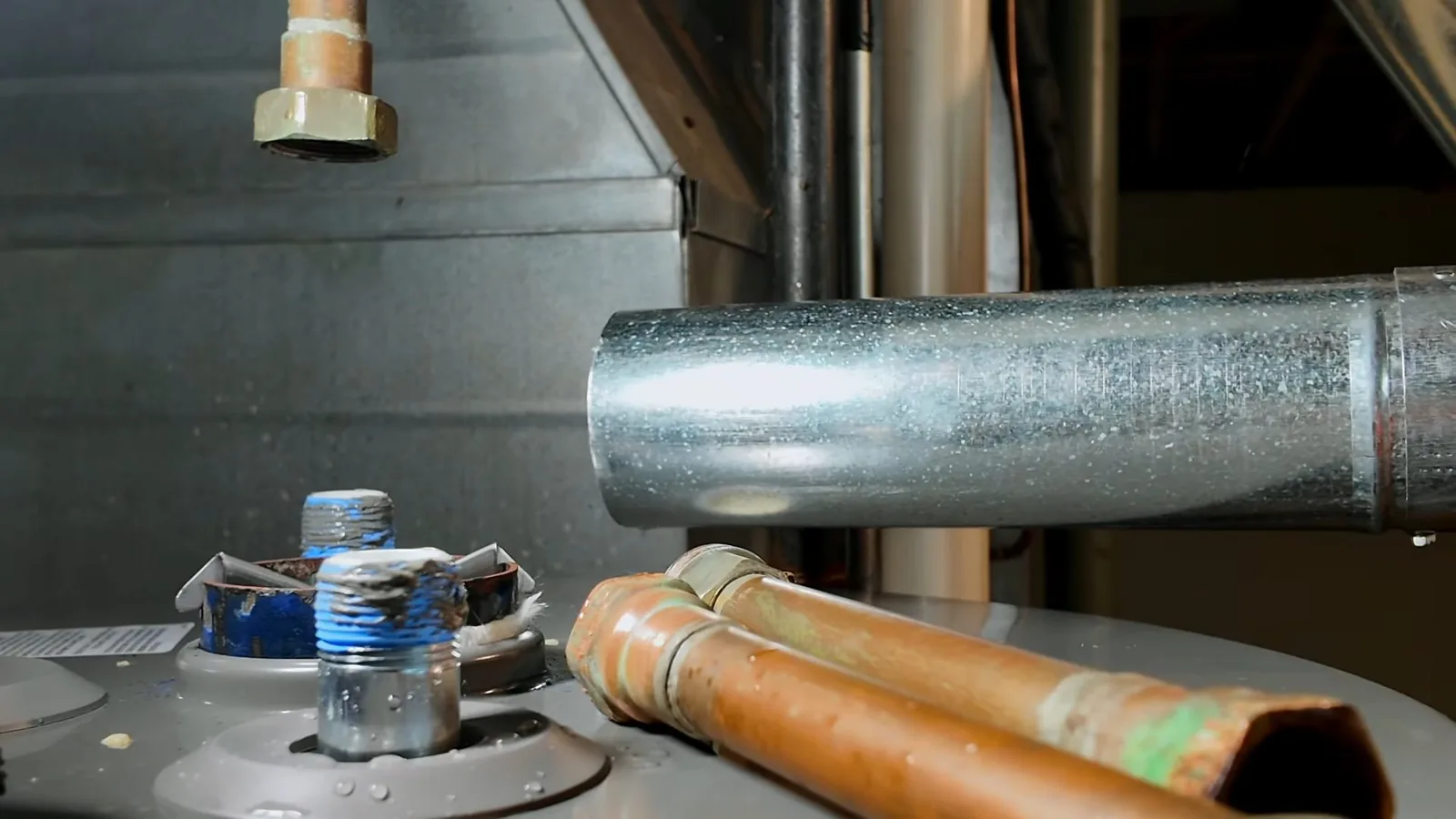 Two copper pipes and a silver metal flue over the top of a tank water heater, with threaded fittings