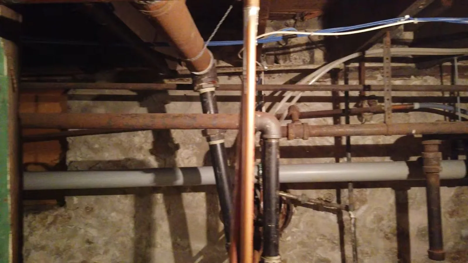 Basement ceiling crowded with old rusted iron, copper and gray plastic pipes running between joists above a stone foundation wall