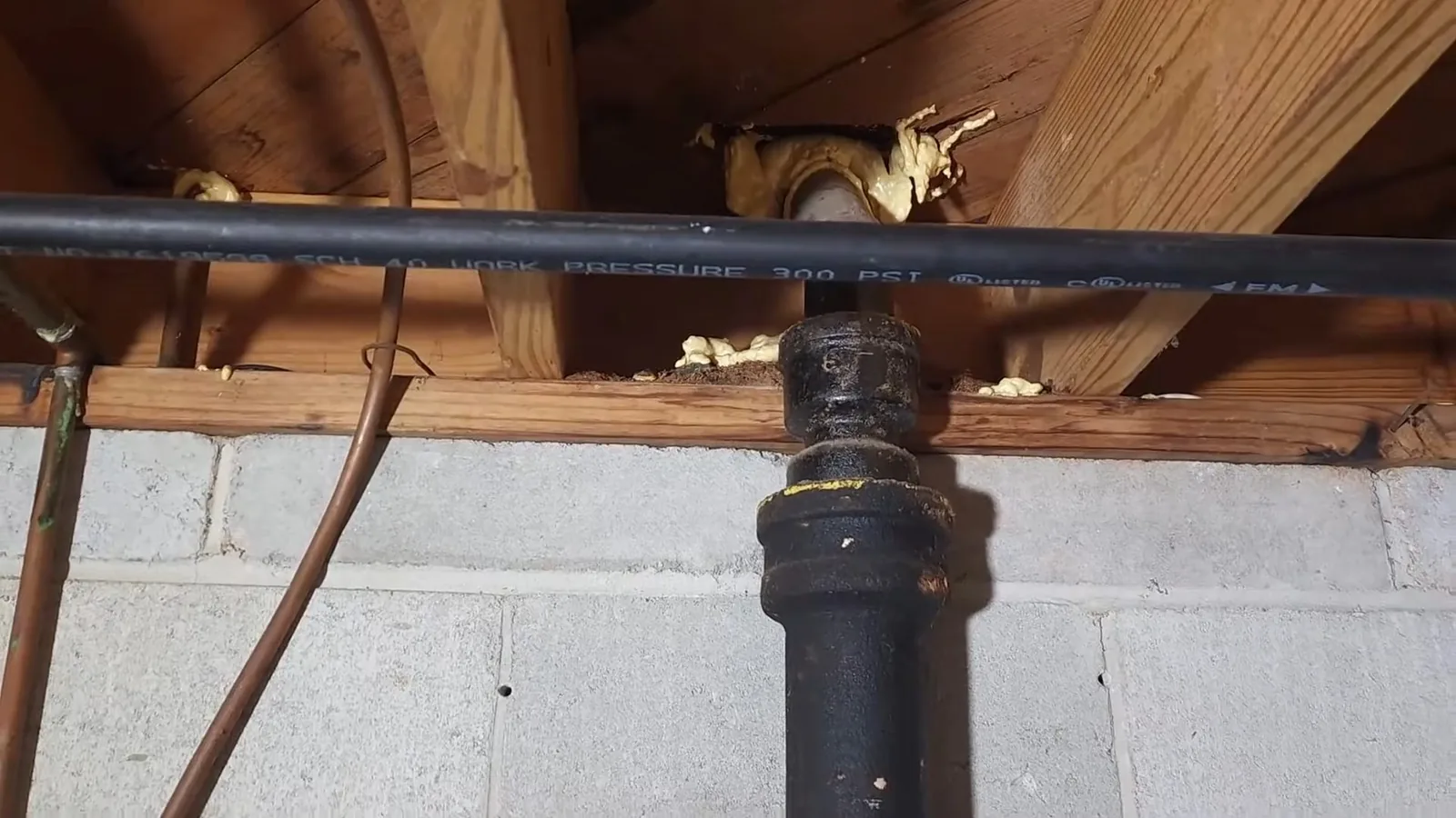 Black cast-iron fitting joined to a long plastic pipe under floor joists, above a concrete block wall
