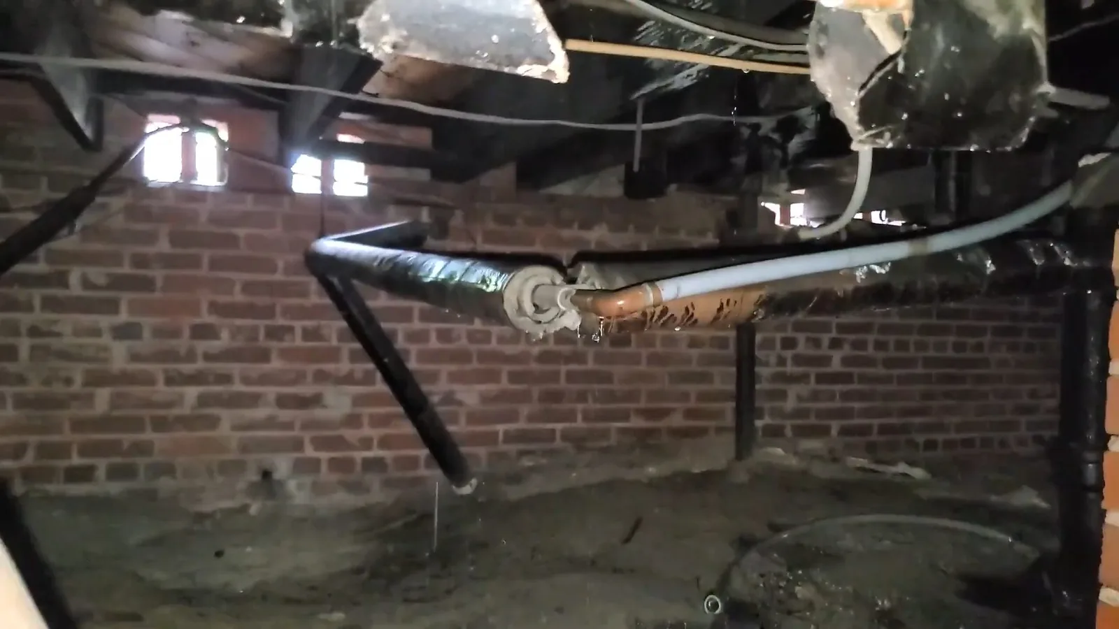 Crawlspace with brick walls, with black wrapped pipes and a white pipe running below the joists above bare soil