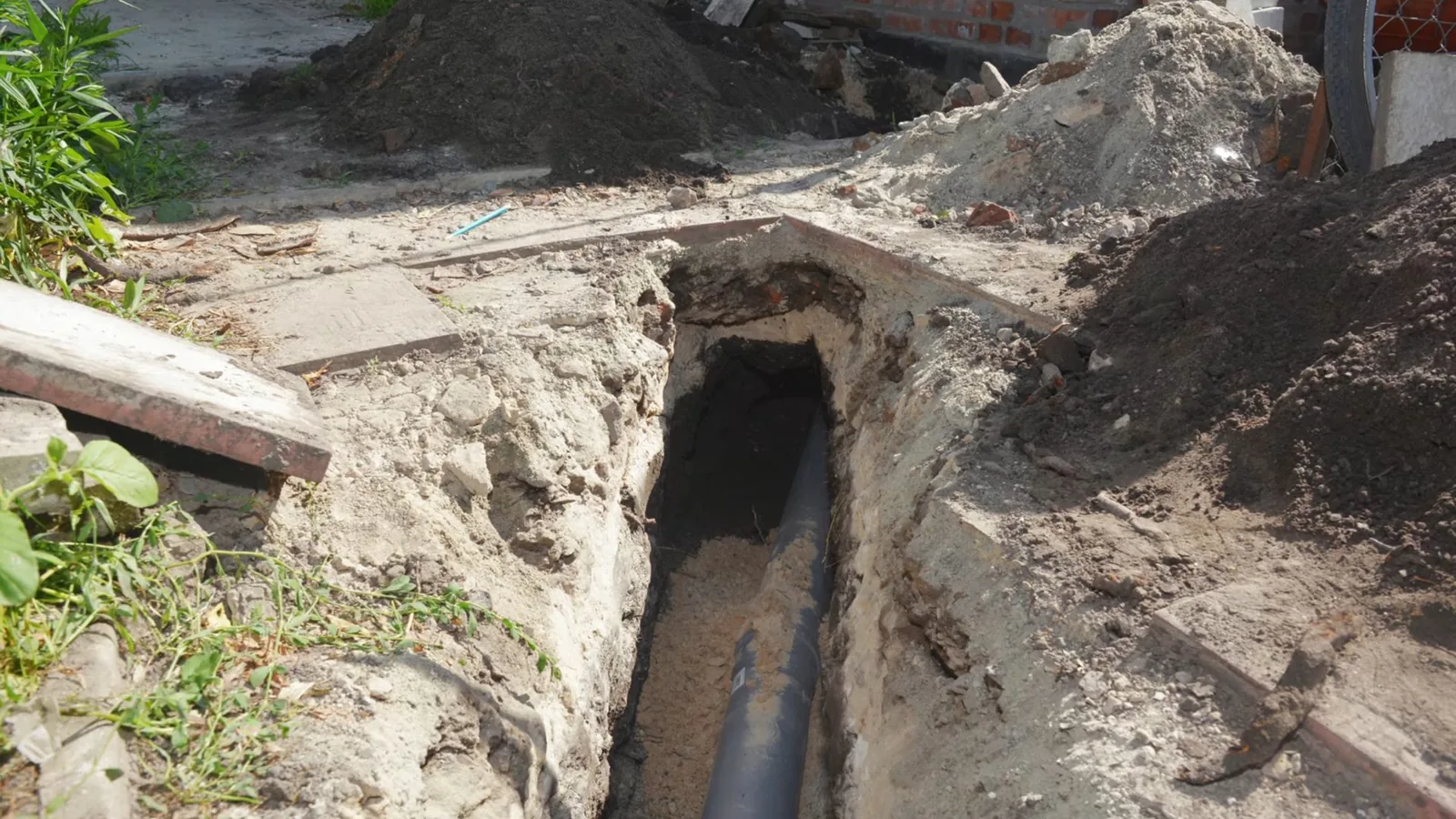 Narrow excavated trench beside a house wall, with a gray sewer pipe visible at the bottom and piles of sandy soil on either side