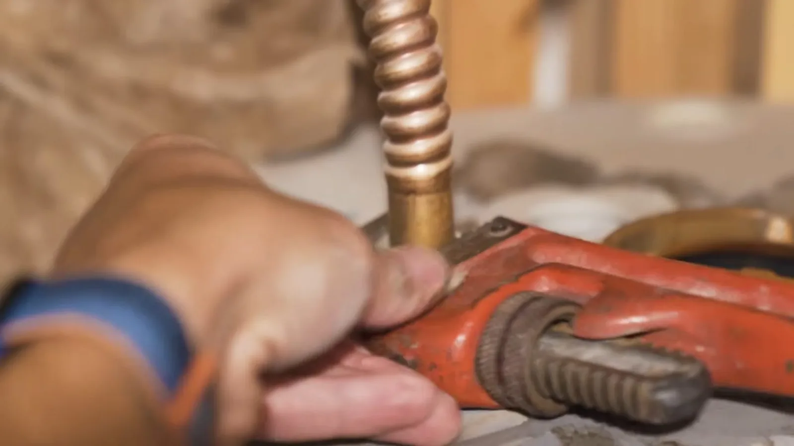 Hand gripping an orange pipe wrench on a brass fitting at the end of a corrugated copper water connector