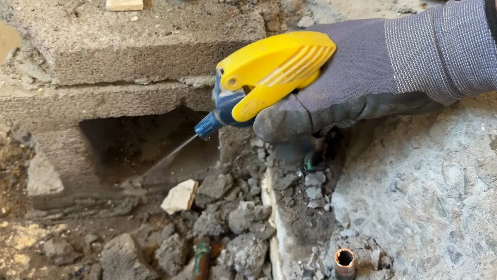 Gloved hand holding a yellow tool at a cut-out in a concrete floor, with a copper pipe visible below