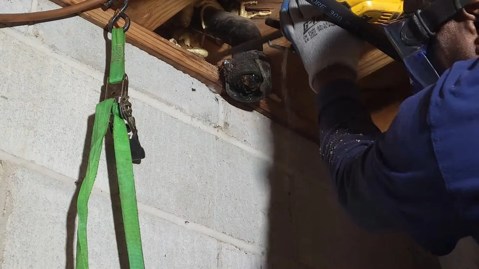 Plumber in a blue work shirt and gloves using a power tool to cut an old cast-iron drain pipe between joists, with a green ratchet strap hanging beside a cinder block wall