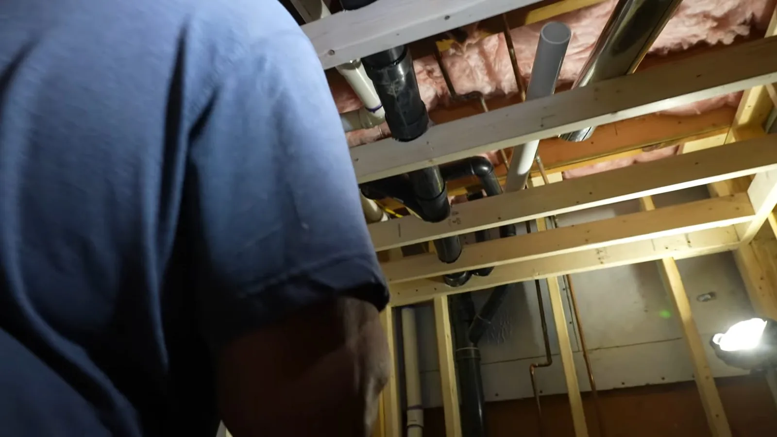 Plumber in a navy shirt working at drain and vent pipes in an unfinished basement ceiling with exposed framing