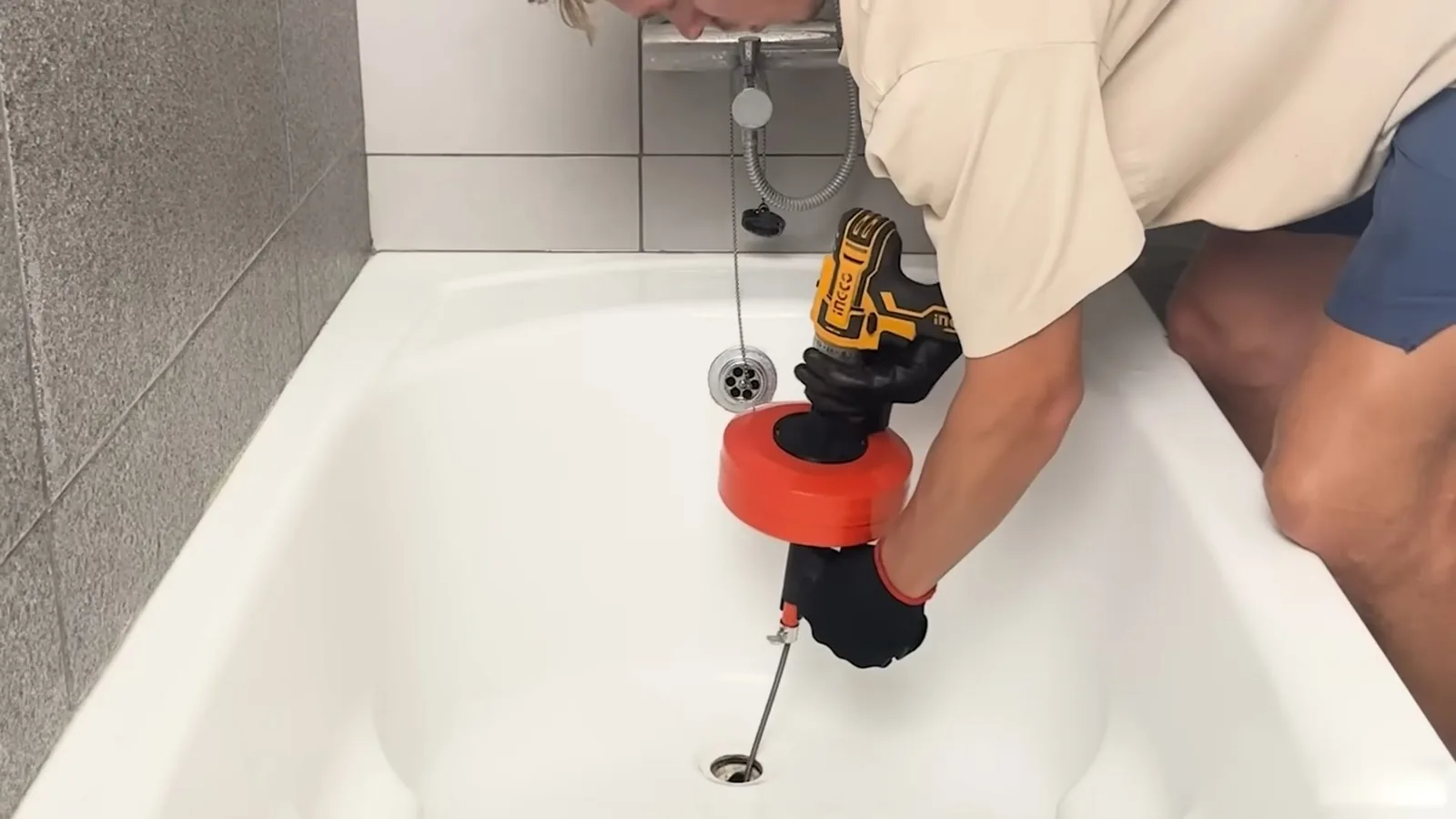 Plumber leaning over a white bathtub, running a cable with a red drum from a cordless drill into the tub drain