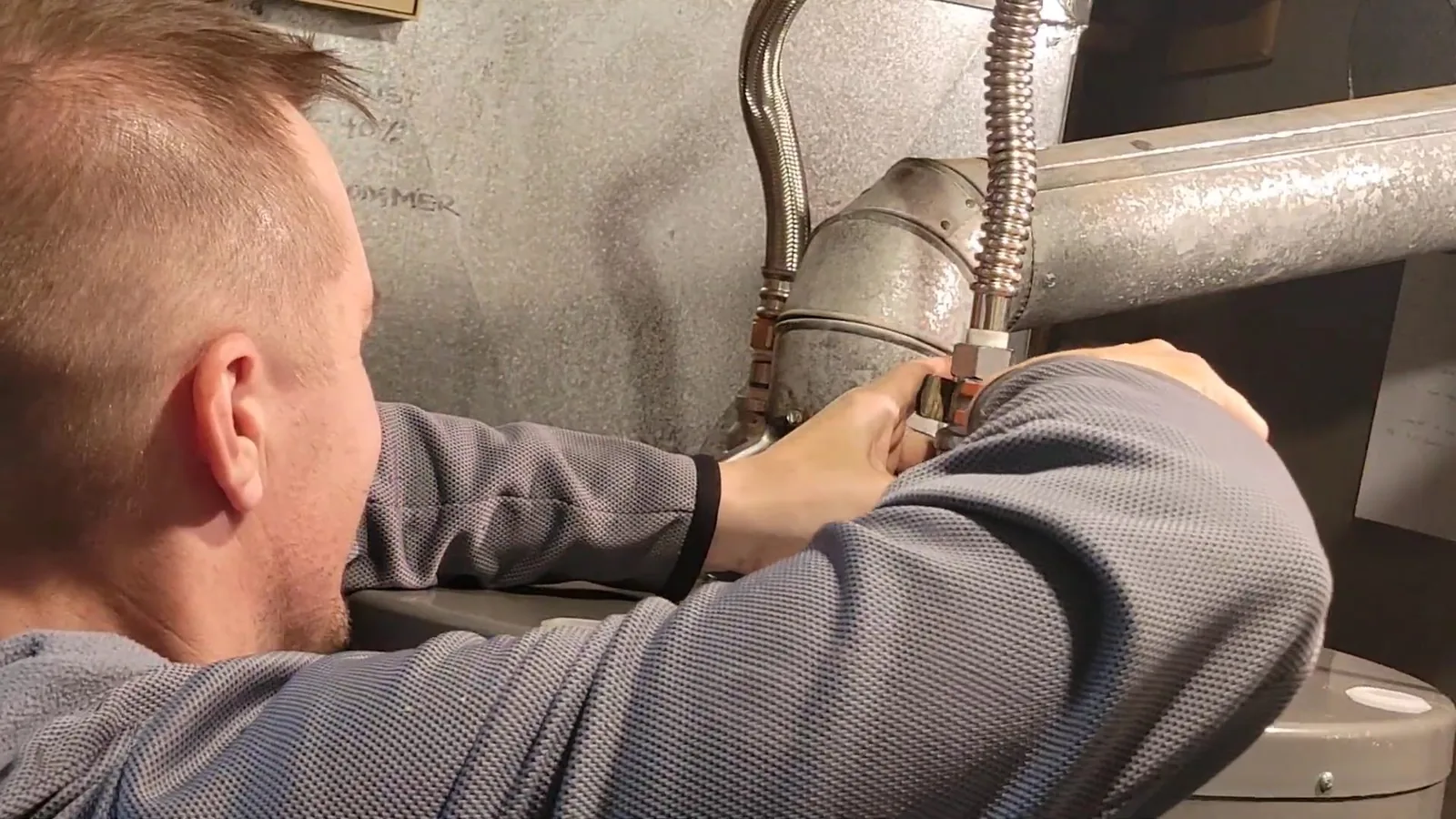 Plumber in a gray shirt leaning in to tighten a braided flexible water line on a tank water heater beside the vent pipe