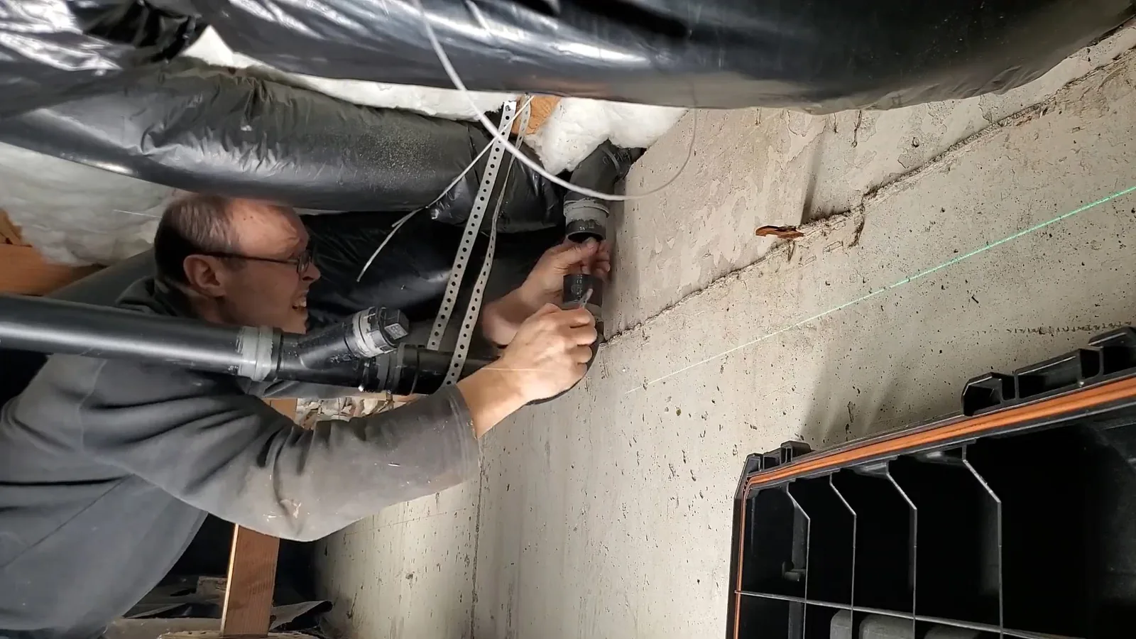 Plumber crouched under floor joists, working on a gray drain pipe fitting, with a green laser line running across the concrete foundation wall
