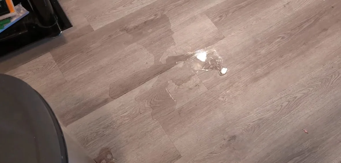 Puddle and wet streaks on a gray wood-look floor, with the edge of an appliance in the corner