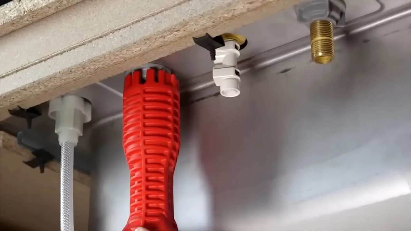 Red basin wrench resting under a sink beside white supply fittings, a hose and a brass connector