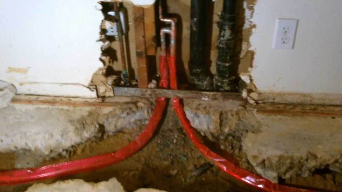 Red flexible supply pipes curving down through a cut in a concrete floor beside an open wall with black drain pipes