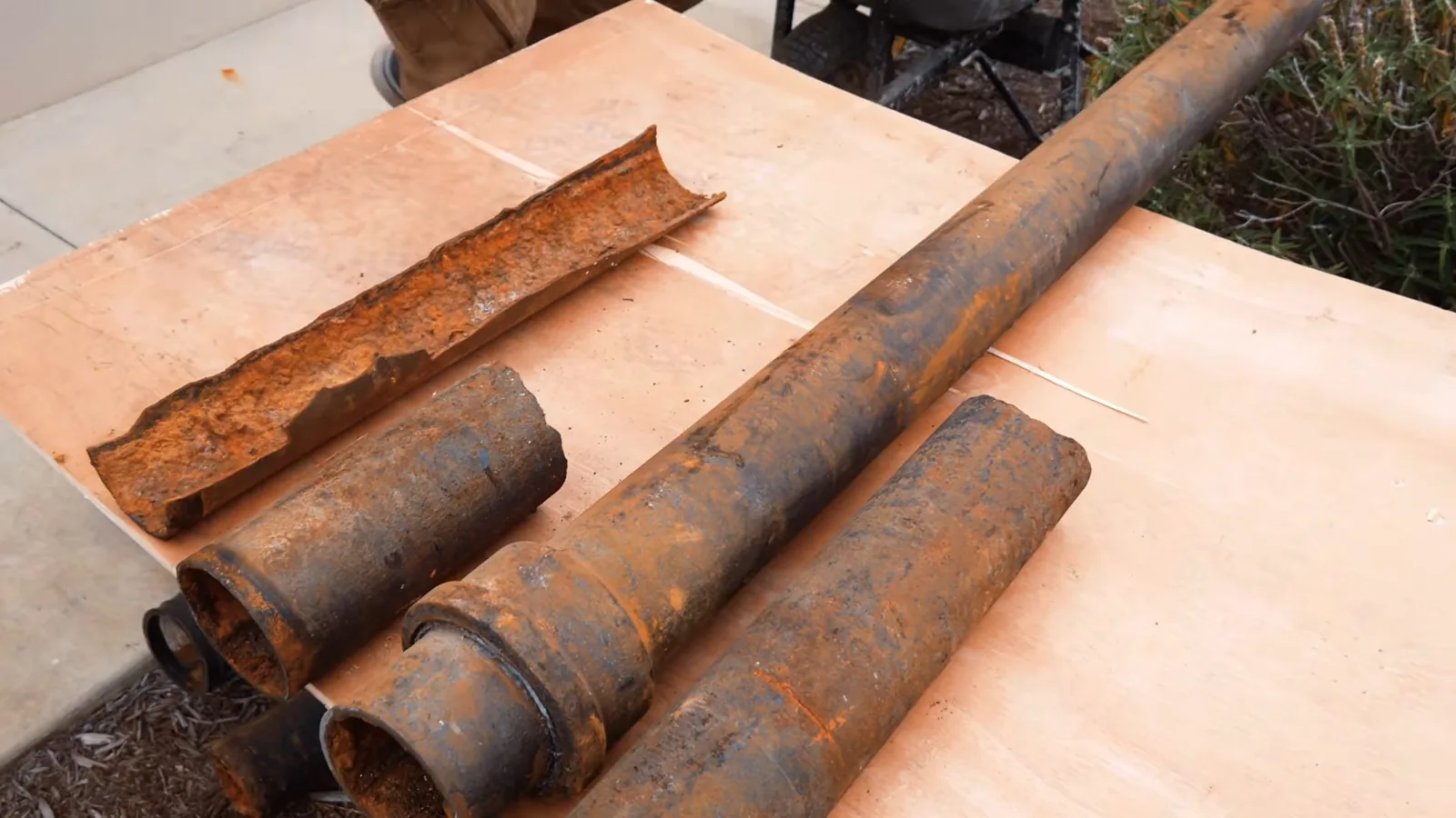 Four sections of rusted cast-iron pipe laid on a plywood sheet, one split lengthwise to show the corroded inside