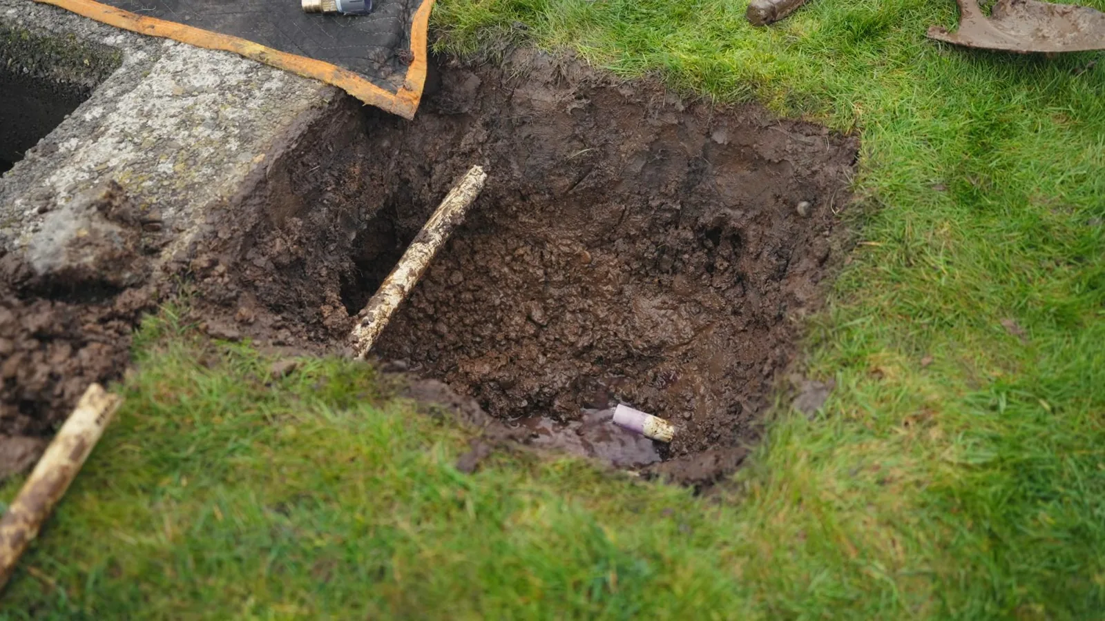 Hole dug in a lawn exposing a buried pipe, with a shovel blade and curb stone at the edge of the grass