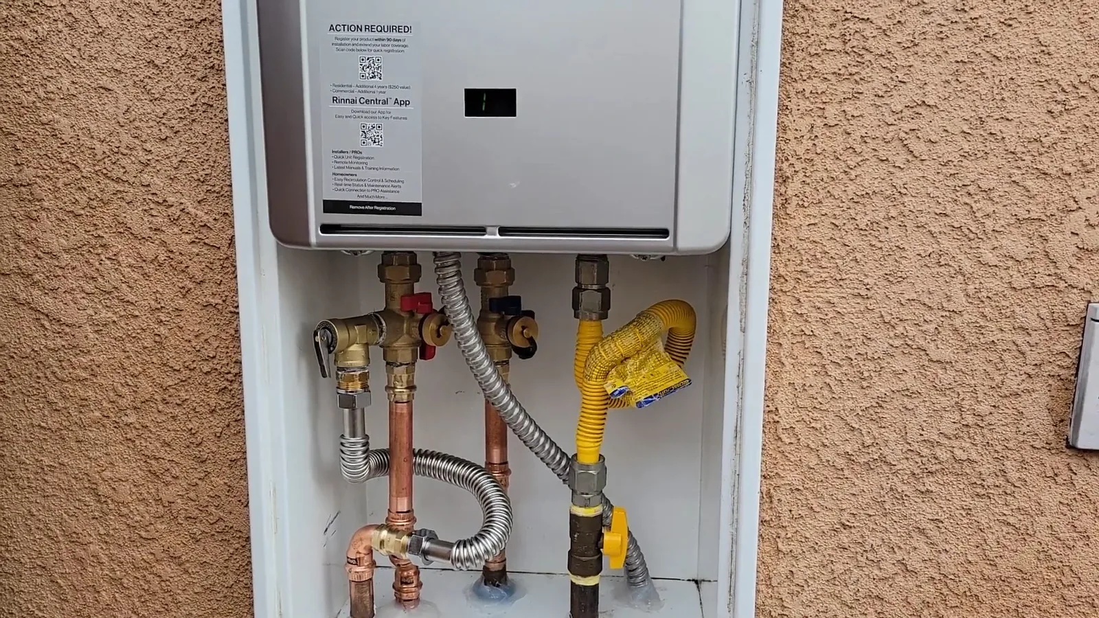 Tankless water heater mounted on a stucco wall, with copper pipes, shutoff valves, a yellow gas line and a stainless flexible hose underneath