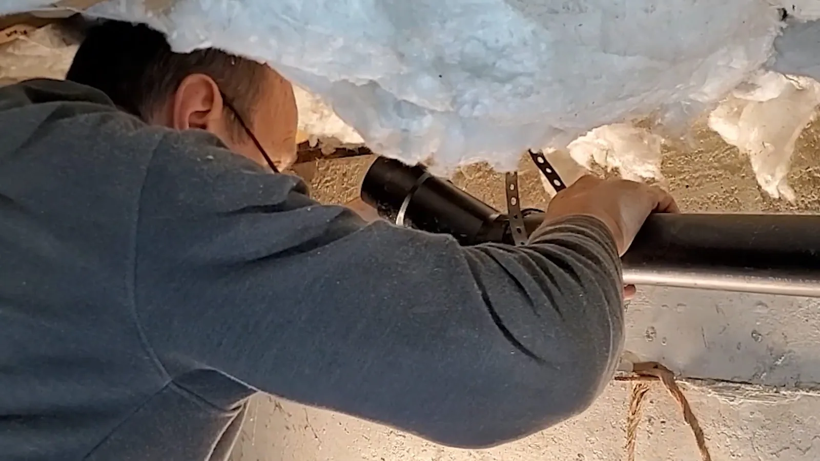 Technician reaching up to a black drain pipe held by a hanger under insulation in a crawlspace