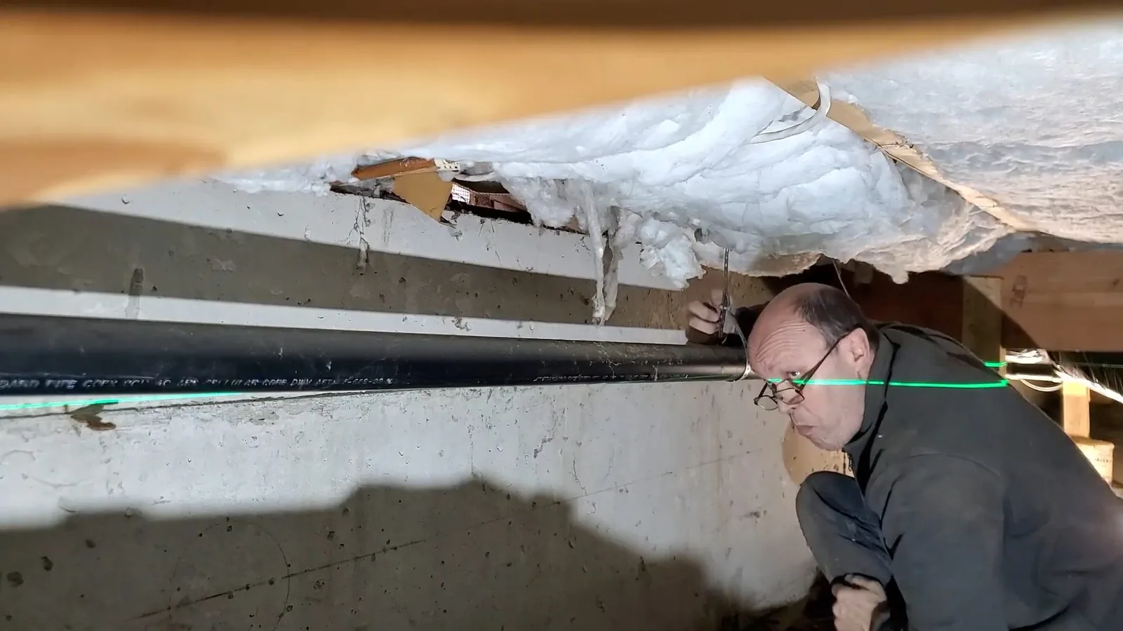 Technician crouched in a crawlspace, looking along a black pipe run that sits below the floor joists